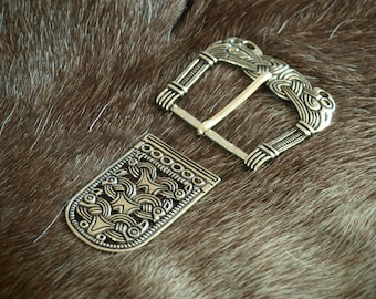 May include: A silver-toned belt buckle with an intricate design. The buckle is made of two pieces, a rectangular plate and a separate buckle. The plate has a decorative design of interlacing lines and circles. The buckle has a similar design with a more prominent central motif.