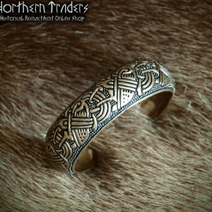 Puede incluir: Un brazalete de metal dorado con intrincados diseños de nudos. El brazalete está sobre un fondo de piel marrón.