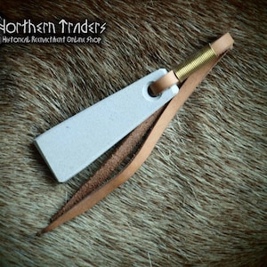 May include: A small, white, rectangular sharpening stone with a leather strap and a brass spring. The stone is attached to the strap with a hole and the spring is attached to the strap with a metal loop. The strap is brown and has a frayed edge. The stone is on a brown, furry surface.