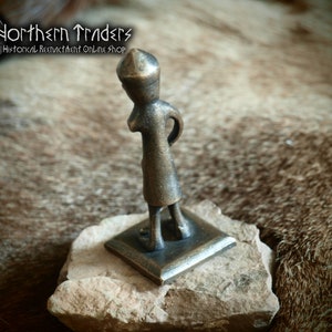 May include: A bronze figurine of a person wearing a pointed hat, standing on a square base. The figurine is on a brown, furry surface.