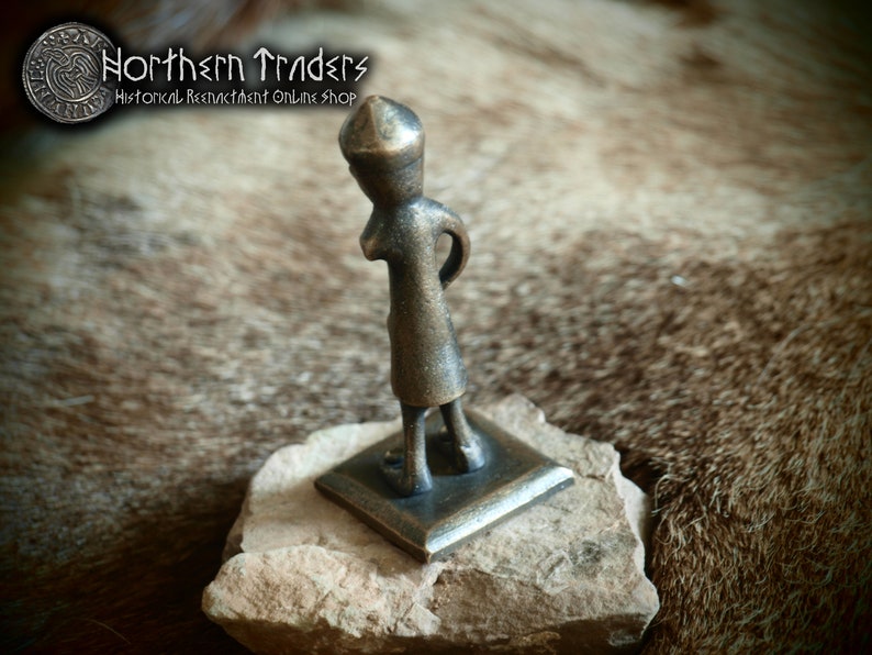 May include: A bronze figurine of a person wearing a pointed hat, standing on a square base. The figurine is on a brown, furry surface.