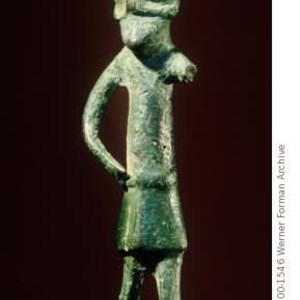 May include: A green bronze figurine of a person wearing a pointed hat. The figurine is standing with its arms at its sides and its legs slightly apart.