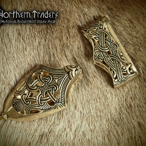 May include: Two antique brass-colored metal belt fittings with intricate Celtic knotwork designs. The fittings are displayed on a textured, light brown surface. The text "Northern Traders Historical Reenactment Online Shop" is visible in the upper left corner.