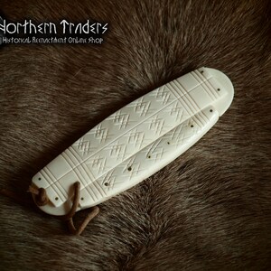 May include: A white bone knife sheath with intricate carvings and a leather strap. The sheath is on a brown fur background.