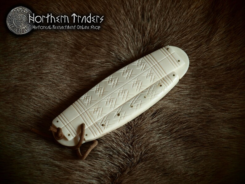 May include: A white bone knife sheath with intricate carvings and a leather strap. The sheath is on a brown fur background.