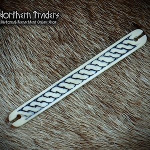 May include: A cream-colored bone artifact with a braided design in dark blue. The item has a slot at each end. The background is a brown, textured surface. The text "Northern Traders Historical Reenactment Online Shop" is visible.