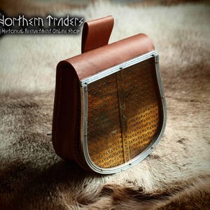 May include: A brown leather pouch with a decorative metal frame and a flap closure. The pouch has a textured front panel and a loop for hanging. The text "Northern Traders Historical Reenactment Online Shop" is visible.