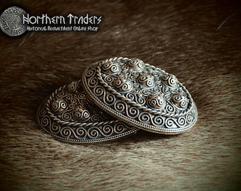May include: Two silver oval-shaped brooches with intricate Celtic knotwork designs. The brooches have a raised, textured surface and are decorated with small, round, raised bumps.