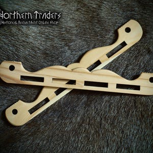 May include: Two light-colored wooden objects with cut-out rectangular sections are crossed over each other. The objects have a curved shape with a hole at each end. The text "Northern Traders Historical Reenactment Online Shop" is in the upper left corner.