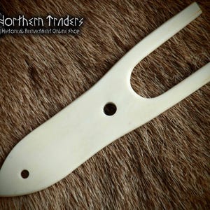 May include: A white bone tool with a handle and two prongs. The tool has two holes in it. The tool is on a brown fur background.