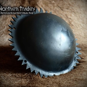Op de afbeelding: Een donkergrijze, ronde metalen schildknop met een glad, koepelvormig midden en een gekartelde, stekelige buitenrand met kleine voorgeboorde gaten. De tekst Northern Traders Historical Reenactment Online Shop is zichtbaar in de linkerbovenhoek.