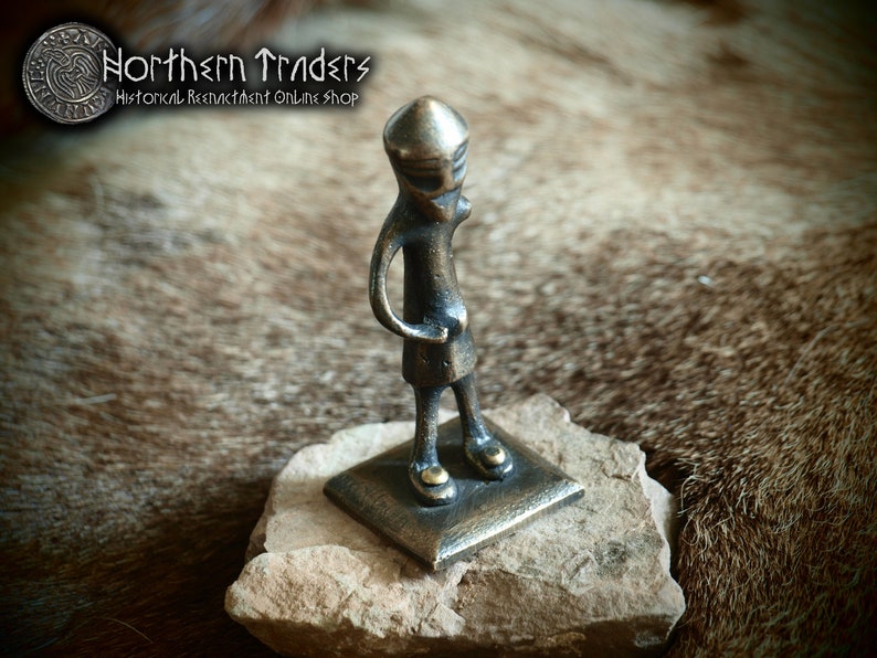 May include: A small bronze figurine of a person wearing a helmet, standing on a square base. The figurine is about 4 inches tall and is made of a dark bronze metal.