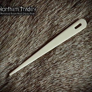 May include: A long, off-white, tapered needle with a single elongated eye at the rounded end. The needle features a smooth, solid surface and a pointed tip. The text Northern Traders Historical Reenactment Online Shop is visible in the corner.