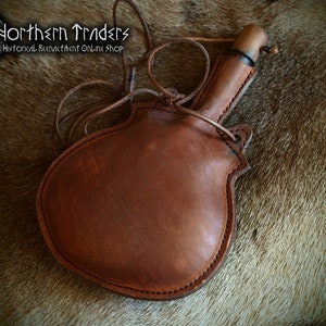 Könnte beinhalten: Eine braune Lederflasche mit einem Holzstopfen und einem Lederriemen. Die Flasche liegt auf einem braunen Fellhintergrund. Northern Traders Historical Reenactment Online Shop.