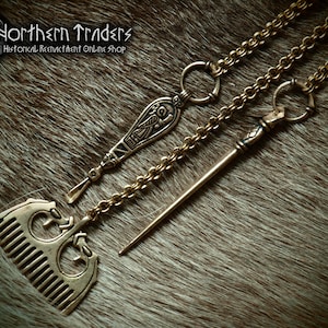 Op de afbeelding: Drie antieke sieraden in Vikingstijl van brons: een kamhanger, een hanger met een gegraveerd vogelontwerp en een lange speld. De stukken zijn verbonden door een ketting en liggen op een bruin, vachtachtig oppervlak. De tekst "Northern Traders Historical Reenactment Online Shop" is inbegrepen.