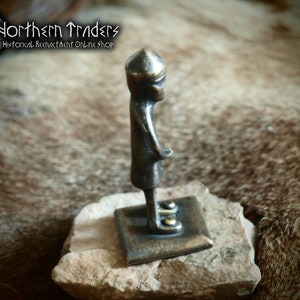 May include: A small bronze figurine of a person wearing a helmet, standing on a square base. The figurine is about 5 inches tall and is made of a dark bronze metal.