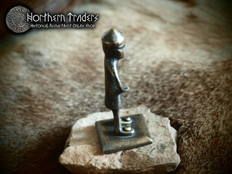 May include: A small bronze figurine of a person wearing a helmet, standing on a square base. The figurine is about 5 inches tall and is made of a dark bronze metal.