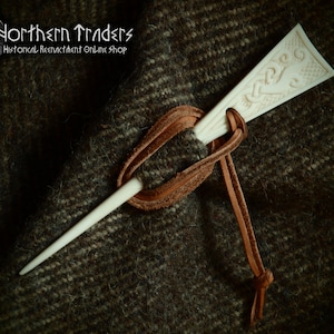 May include: A white bone pin with a carved design on the top. The pin is tied to a brown leather cord.