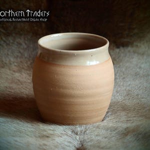 Peut inclure: Un mug en céramique fait à la main, de forme arrondie, avec un extérieur beige clair et un bord beige plus clair. Le mug a un aspect rustique et texturé, sur un fond flou. Le logo "Northern Traders" est visible en haut à gauche.