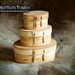 Könnte beinhalten: Ein Stapel aus drei ovalen Holzkisten mit hellen und dunklen Holzakzenten. Jede Kiste hat einen Klappdeckel und einen dekorativen Metallverschluss. Die Kisten sind auf einer strukturierten, braunen Oberfläche ausgestellt.