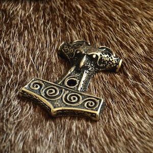 Pode incluir: Um pingente de metal dourado em forma de martelo com intrincados desenhos em espiral. O pingente está sobre uma superfície peluda marrom.