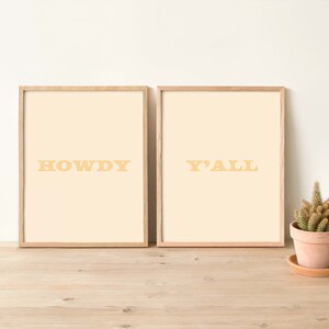 May include: Two framed prints with a light beige background and a light brown wood frame. The print on the left says "HOWDY" in yellow letters. The print on the right says "Y'ALL" in yellow letters.
