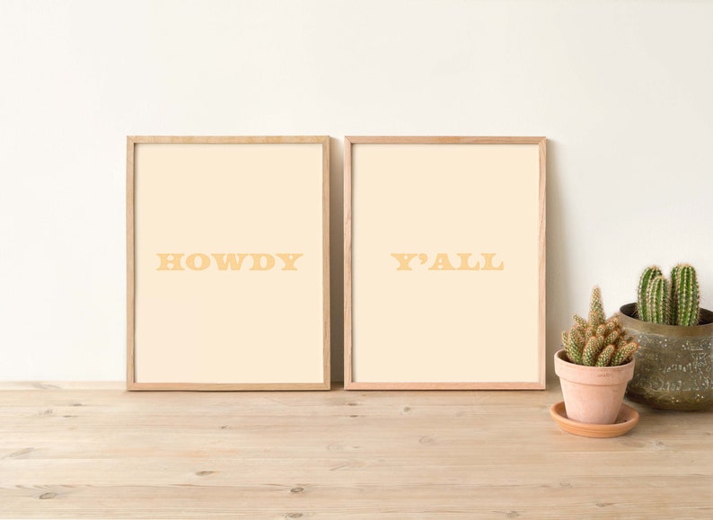 May include: Two framed prints with a light beige background and a light brown wood frame. The print on the left says "HOWDY" in yellow letters. The print on the right says "Y'ALL" in yellow letters.