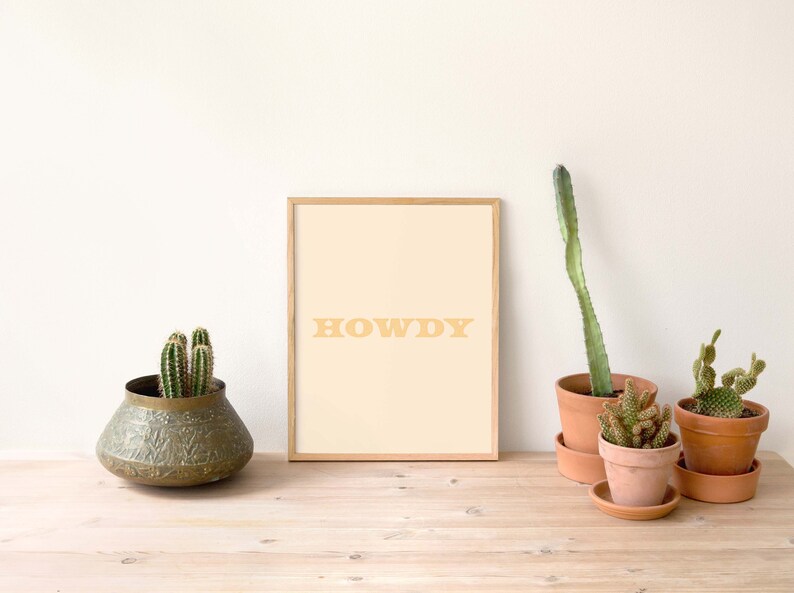 May include: A framed print with a light beige background and the word "HOWDY" in a light orange color. The print is sitting on a wooden table with several potted cacti.