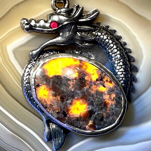 May include: A silver dragon pendant with a red eye and an oval-shaped stone. The stone features orange, yellow, and black hues. The pendant is set against a light brown and white patterned background.