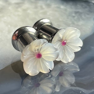 Puede incluir: Un par de dilatadores de oreja de plata con diseños de flores blancas y centros de gemas rosas.