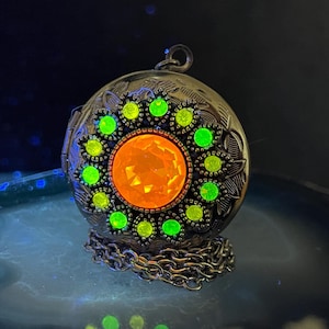 May include: A silver locket necklace with a large orange and green gem in the center. The locket is surrounded by a circle of smaller green gems.