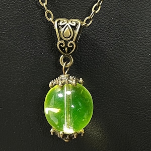 Puede incluir: Un collar con un colgante de esfera de vidrio verde y una cadena y cierre de bronce ornamentado.