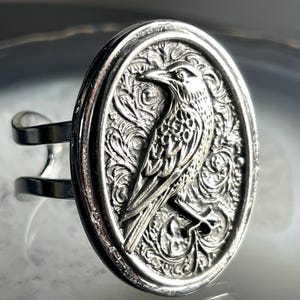 May include: Silver-toned ring featuring an oval centerpiece with a detailed raven design. The raven is surrounded by ornate, swirling patterns. The ring has a split band design for an adjustable fit. The ring is made of a shiny, metallic material.