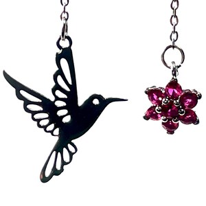May include: A pair of drop earrings. One earring has a black hummingbird outline, the other a cluster of pink gemstones in a flower shape. Both are suspended from delicate silver chains.