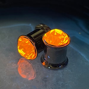 May include: Two black ear gauges with bright orange, faceted gemstone tops. The plugs are cylindrical with a polished finish, reflecting the light. The gemstones have a brilliant, multi-faceted cut, creating a sparkling effect.
