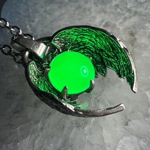 K&ouml;nnte beinhalten: Eine silberne Halskette mit einem Anh&auml;nger, der ein Paar Fl&uuml;gel zeigt, die eine leuchtend gr&uuml;ne Kugel halten.