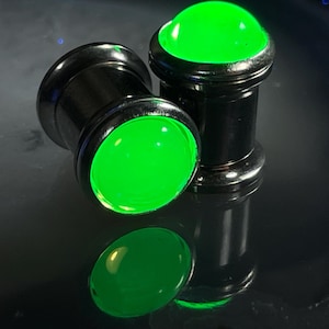 May include: Two black metal ear gauges with green glass inserts. The gauges are cylindrical and have a smooth, polished finish.