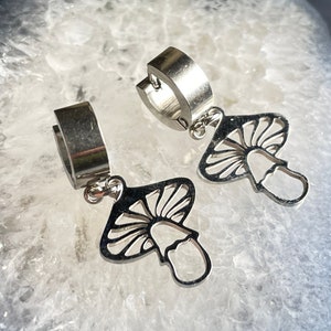 May include: A pair of silver-colored hoop earrings. Each earring features a mushroom charm with a detailed, openwork design. The earrings are displayed on a white and gray surface.