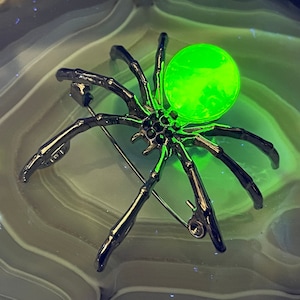 May include: A dark metal spider brooch with a glowing green orb on its back. The spider has detailed legs and a pin closure. The brooch is set against a light green and white background.
