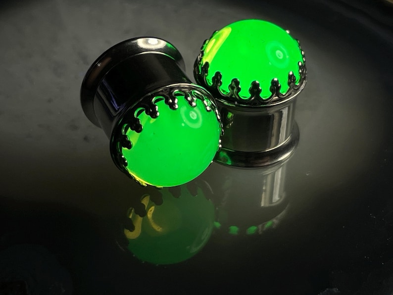 May include: Two green glow-in-the-dark ear gauges with silver metal accents. The gauges have a decorative crown design around the green glass.