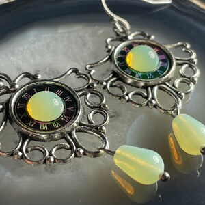 May include: Silver earrings with a clock face design and a pale green stone. The clock face has Roman numerals and a iridescent background. The earrings have a dangling pale green stone.