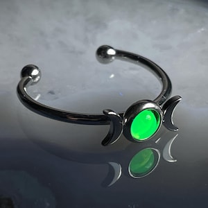 May include: A silver-toned nose ring featuring a bright green center stone. The ring has a curved design with a small ball on each end. The center stone is flanked by two crescent moon shapes.