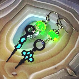 May include: A pair of dangle earrings featuring black key-shaped charms with teal accents. Each earring has a bright green, glowing orb and silver-colored ear wires. The earrings are set against a marbled, neutral-toned background.