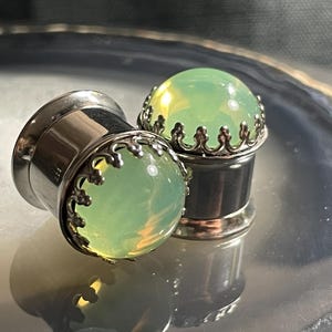 May include: Two silver ear gauges with green opal stone inserts. The gauges have a decorative silver trim around the stone.