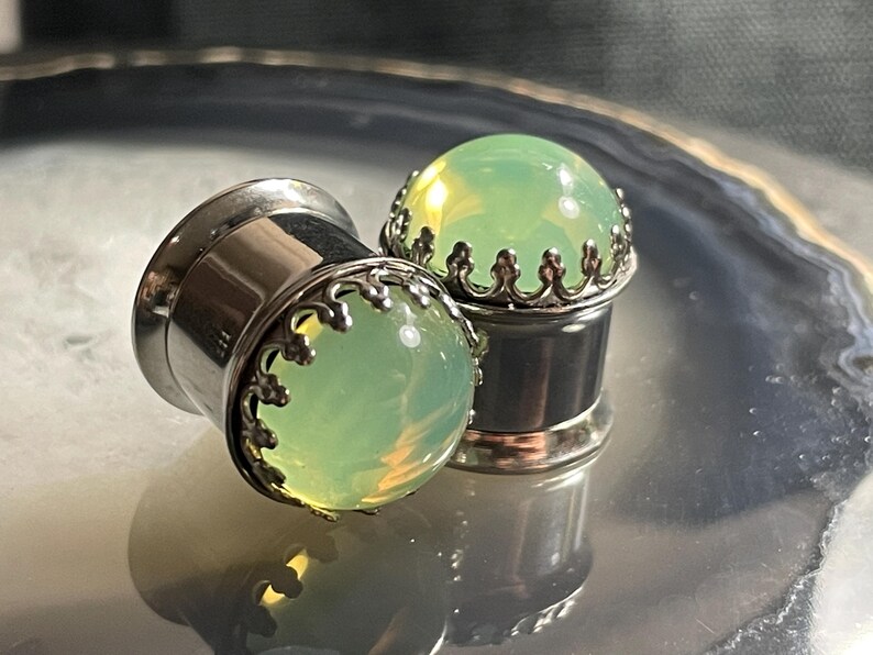 May include: Two silver ear gauges with green opal stone inserts. The gauges have a decorative silver trim around the stone.