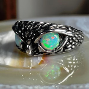 Könnte beinhalten: Ein silberner Ring mit einem Eulen-Design. Die Augen der Eule sind große, runde Opale mit grünen, blauen und rosa Farbtönen. Das Ringband ist strukturiert, um Federn nachzuahmen, wodurch ein detailliertes und einzigartiges Schmuckstück entsteht.