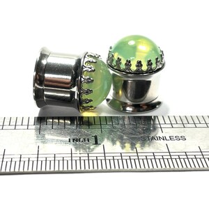 May include: Two silver ear gauges with green glass inserts and a silver crown design. The gauges are measured against a ruler.