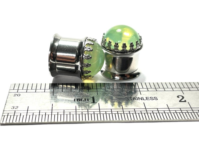 May include: Two silver ear gauges with green glass inserts and a silver crown design. The gauges are measured against a ruler.