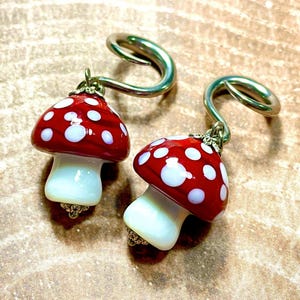 May include: A pair of ear gauges featuring dangling glass mushroom charms. Each mushroom has a glossy red cap with white polka dots and a smooth white stem. The charms are attached to silver-toned metal spiral ear plugs with small metal loops.