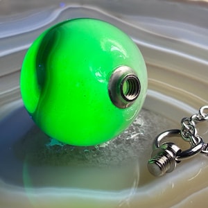 May include: A spherical pendant featuring a vibrant neon green marbled finish with a metallic threaded opening. A separate silver-toned metal screw component is attached to a matching link chain, designed to fit into the pendant's threaded aperture.
